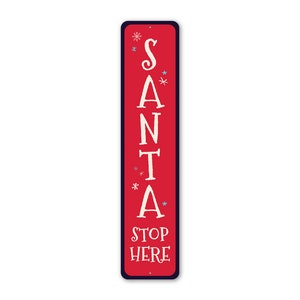 May include: Red metal sign with white text that reads "SANTA STOP HERE" with snowflakes in blue and white around the word "SANTA".