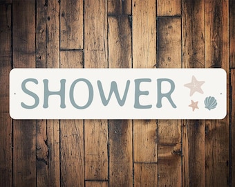 May include: A rectangular, off-white sign with the word "SHOWER" in blue-grey letters. The sign features a starfish and seashell graphic. The background is a wooden wall.