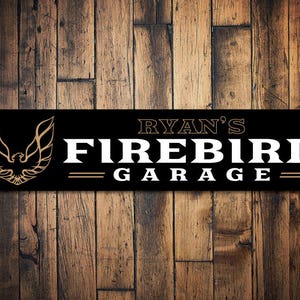 May include: Black metal sign with gold lettering that reads "Ryan's Firebird Garage". A gold bird logo is on the left side of the sign.