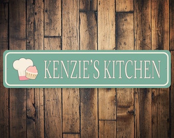 May include: A teal blue metal sign with white text that reads "KENZIE'S KITCHEN". A white chef's hat and a pink cupcake are on the left side of the sign.