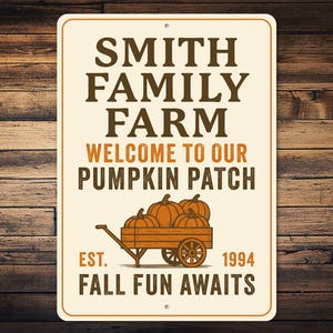 May include: Cream-coloured rectangular sign with brown and orange text: "SMITH FAMILY FARM WELCOME TO OUR PUMPKIN PATCH". A wooden cart carries pumpkins. The sign also reads "EST. 1994 FALL FUN AWAITS".