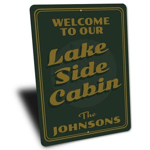 May include: A rectangular, dark green metal sign with a gold border and text. The sign reads "WELCOME TO OUR LAKE SIDE CABIN" in gold lettering, with "The Johnsons" below. The sign is angled slightly.