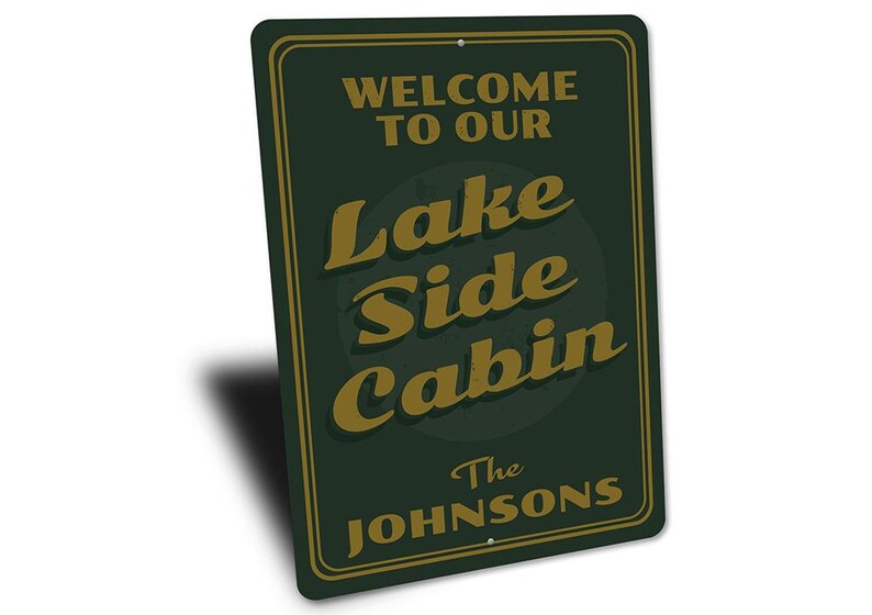 May include: A rectangular, dark green metal sign with a gold border and text. The sign reads "WELCOME TO OUR LAKE SIDE CABIN" in gold lettering, with "The Johnsons" below. The sign is angled slightly.