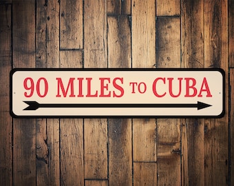 May include: A white metal sign with black trim and red text that reads "90 MILES TO CUBA".  Two black arrows point in opposite directions.