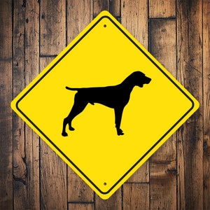 May include: Yellow diamond-shaped warning sign with a black silhouette of a dog. The sign has a hole at the top for hanging.