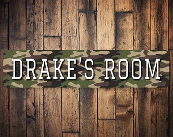 Puede incluir: Un letrero rectangular con un patrón de camuflaje en tonos de verde, marrón y beige. El letrero tiene texto blanco que dice "DRAKE'S ROOM".