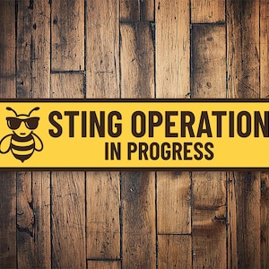 May include: Yellow sign with black text and a cartoon bee wearing sunglasses. The sign reads "Sting Operation in Progress".