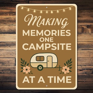 May include: A rectangular brown metal sign with the words "Making Memories One Campsite At A Time" in white and cream lettering. A cream-colored camper illustration is centered on the sign, with floral accents.