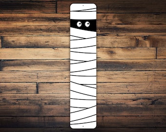 May include: A white mummy with black eyes peeking out from the bandages. The mummy is on a brown wood background.