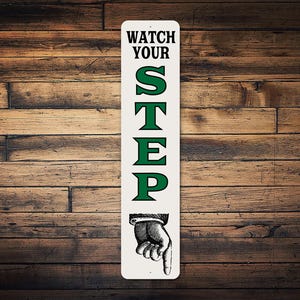 May include: A rectangular white metal sign with the words "WATCH YOUR STEP" in black and green. The sign features a drawing of a hand pointing down. The background is a wooden surface.