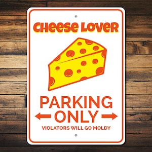 May include: White metal sign with orange text that reads "Cheese Lover Parking Only" with an arrow pointing left and right.  A yellow wedge of cheese with orange spots is above the text.  The text "Violators Will Go Moldy" is below the arrows.