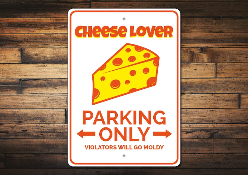 May include: White metal sign with orange text that reads "Cheese Lover Parking Only" with an arrow pointing left and right.  A yellow wedge of cheese with orange spots is above the text.  The text "Violators Will Go Moldy" is below the arrows.