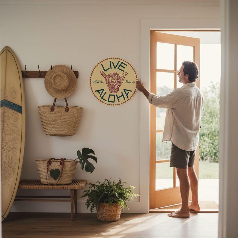 A man is standing in a room with a surfboard on the wall, a round wall sign that says "LIVE ALOHA", and a potted plant.