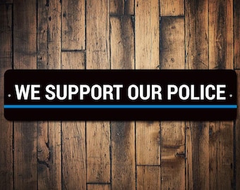 May include: Black and white sign with a blue stripe across the middle. The sign reads "WE SUPPORT OUR POLICE."