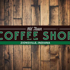 May include: A rectangular metal sign for "Old Town Coffee Shop" in Zionsville, Indiana. The sign has a dark brown background with green and white lettering. The sign is mounted on a wooden plank wall.