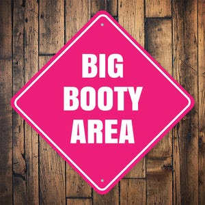 Op de afbeelding: Een roze ruitvormig bord met witte tekst die "BIG BOOTY AREA" zegt op een houten achtergrond.