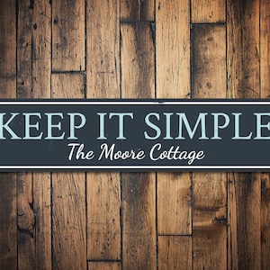 May include: A black and white sign with the text "KEEP IT SIMPLE" in blue lettering. Below the text is "The Moore Cottage" in white lettering.