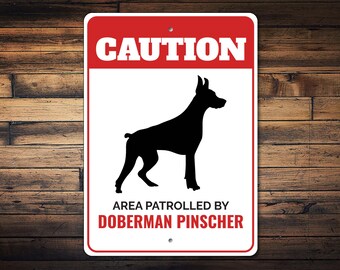 May include: A white and red sign with the word "CAUTION" in large white letters. A black silhouette of a Doberman Pinscher is in the center of the sign. The text "AREA PATROLLED BY DOBERMAN PINSCHER" is at the bottom of the sign.