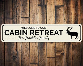 May include: White rectangular sign with black text and a silhouette of a deer. The sign reads "Welcome to our Cabin Retreat The Franklin Family".