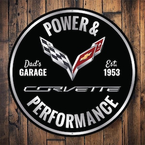 May include: A round black and silver sign with the text "POWER & PERFORMANCE" and "CORVETTE" in silver. The sign also includes the text "Dad's GARAGE" and "Est. 1953". The background is a wooden surface.