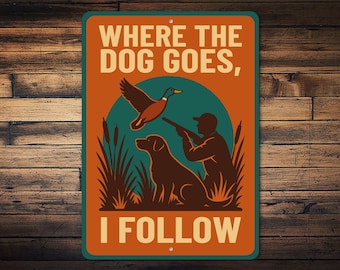 Op de afbeelding: Rechthoekig metalen bord met een jachtthema. Het bord toont een illustratie van een jager, hond en eend in bruin, oranje en blauwgroen. De tekst luidt "WHERE THE DOG GOES, I FOLLOW."