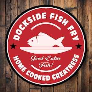 May include: Red and white circular sign with the text "Dockside Fish Fry Home Cooked Greatness" and a white silhouette of a fish on a plate. The text "Good Eatin Fish!" is inside the circle.
