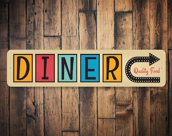 Puede incluir: Un letrero de metal de estilo vintage con la palabra "DINER" en letras mayúsculas de colores. El letrero tiene una flecha negra que apunta hacia la derecha con el texto "Quality Food" en rojo.