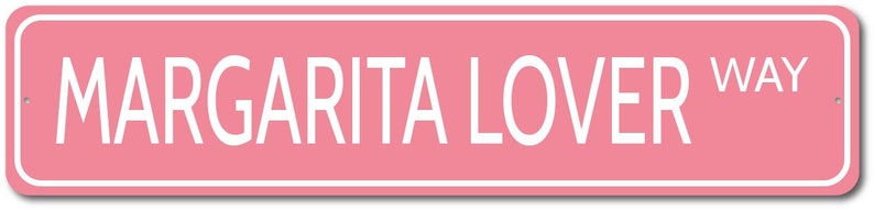 May include: Pink rectangular sign with white border and white text that reads "MARGARITA LOVER WAY".