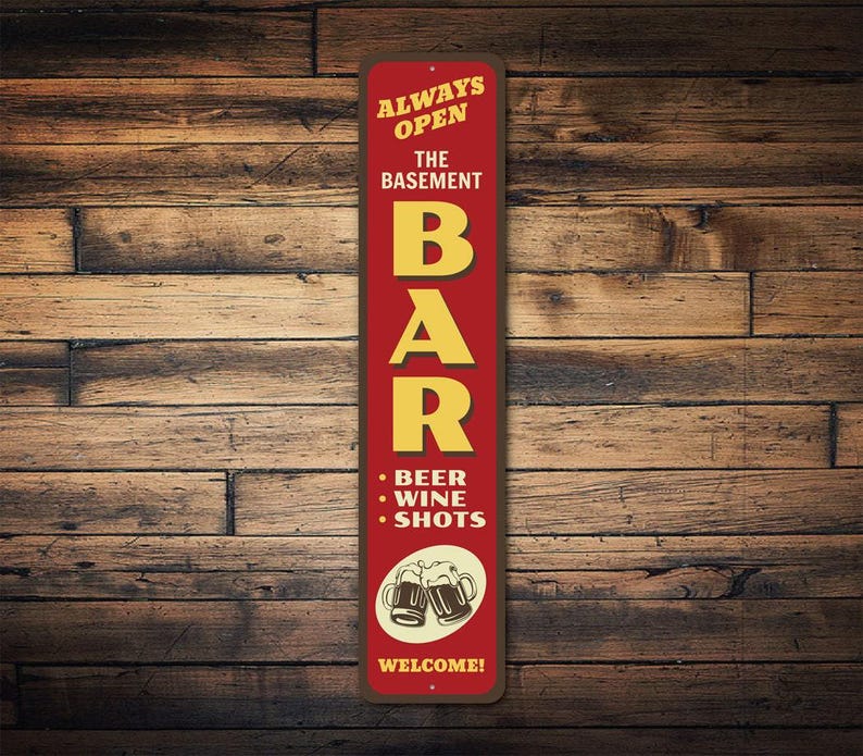 Może przedstawiać: Czerwono-ż&oacute;łty znak z napisem "Always Open The Basement Bar Beer Wine Shots Welcome!" i grafiką dw&oacute;ch kufli piwa.