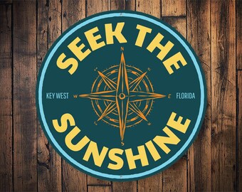 May include: A round, blue and yellow sign with a compass rose in the center. The sign says "Seek the Sunshine" with "Key West" and "Florida" on either side of the compass rose.