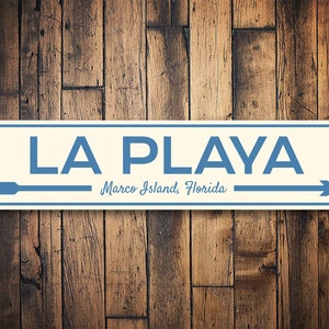 May include: A white metal sign with blue text and arrows that reads "LA PLAYA Marco Island, Florida".