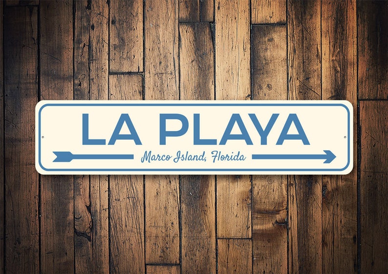 May include: A white metal sign with blue text and arrows that reads "LA PLAYA Marco Island, Florida".