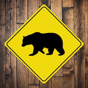 May include: Yellow diamond-shaped warning sign with a black silhouette of a bear walking. The sign is on a wooden background.