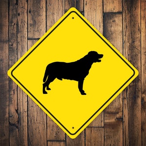 May include: A yellow diamond-shaped warning sign with a black silhouette of a dog inside. The sign is made of metal and has a hole at the top for hanging.