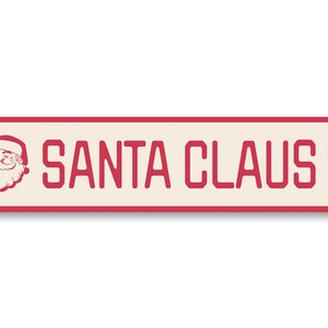 Op de afbeelding: Een rood en wit metalen bord met de tekst "SANTA CLAUS LN" en een cartoonafbeelding van de kerstman in de linkerbovenhoek.