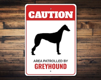 Può includere: Cartello metallico rettangolare con bordo rosso e la scritta "CAUTION" in lettere bianche nella parte superiore. Sotto, una silhouette nera di un levriero è centrata su uno sfondo bianco. Il testo "AREA PATROLLED BY GREYHOUND" è in basso.
