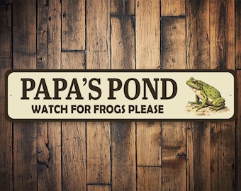 Może przedstawiać: Metalowy znak w kolorze brązowym i białym z napisem "Papa's Pond" i "Watch for Frogs Please". Po prawej stronie znaku znajduje się zielona żaba.