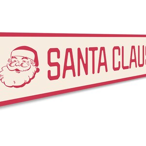 Op de afbeelding: Een rood en wit metalen bord met een afbeelding van de kerstman en de tekst "SANTA CLAUS LN".
