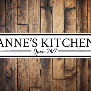 May include: White rectangular sign with black text that reads "Anne's Kitchen - Open 24/7". The sign is on a wood background.