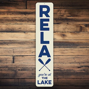 Puede incluir: Un letrero de metal blanco con texto azul que dice "RELAX you're at THE LAKE". El letrero tiene un par de remos azules cruzados en la parte superior.