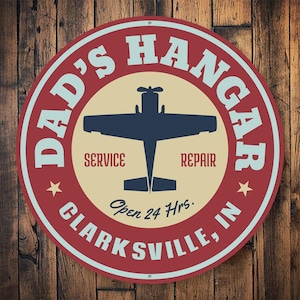 May include: Red and white round sign with the text "Dad's Hangar" in a white circle with a blue silhouette of a small airplane inside. The text "Service" and "Repair" are below the airplane. The text "Open 24 Hrs." is below the airplane and the text "Clarksville, IN" is below that.