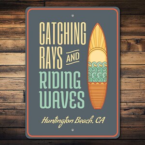 May include: Rectangular metal sign with the words "Catching Rays and Riding Waves" and a surfboard illustration. The sign is dark gray with an orange border and the text "Huntington Beach, CA" at the bottom. The background is a wooden wall.