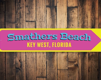 Può includere: Un cartello in metallo giallo e rosa con una freccia blu che punta a destra. Il cartello recita "Smathers Beach KEY WEST, FLORIDA".