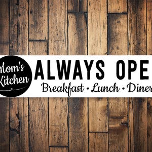 May include: A rectangular white sign with black text and a wooden background. The sign reads "Mom's Kitchen ALWAYS OPEN Breakfast • Lunch • Diner" in a mix of fonts. The words "Mom's Kitchen" are in a black circle.
