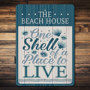 Puede incluir: Un letrero rectangular de madera con borde azul y centro blanco. El letrero dice "THE BEACH HOUSE" en la parte superior y "One Shell of a Place to LIVE" en una fuente decorativa. Acentos florales están dispersos alrededor del texto.