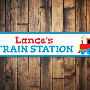 May include: A blue and white sign with the text "Lance's Train Station" in red and blue letters. A red toy train with black wheels and a green and yellow smokestack is on the right side of the sign.