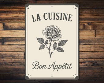 May include: A rectangular sign with a cream background and a black border. The sign features the words "LA CUISINE" above a detailed rose illustration and the words "Bon Appétit" below. The sign is mounted on a wooden background.