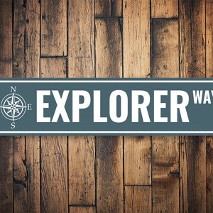 May include: A gray metal sign with white text that reads "EXPLORER WAY". A compass with the directions N, S, E, and W is on the left side of the sign.