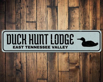 Może przedstawiać: Prostokątny znak z napisem "DUCK HUNT LODGE" w kolorze czarnym, z napisem "EAST TENNESSEE VALLEY" poniżej. Czarna sylwetka kaczki znajduje się po prawej stronie. Znak jest umieszczony na drewnianym tle.