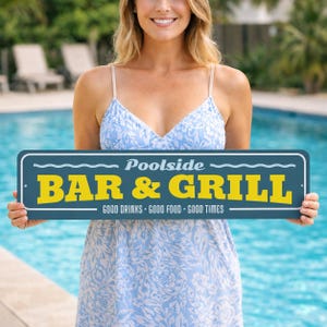 May include: A rectangular sign with the words "Poolside BAR & GRILL" in yellow letters on a dark blue background. The sign also includes the text "GOOD DRINKS - GOOD FOOD - GOOD TIMES" in white. The sign is held in front of a pool.